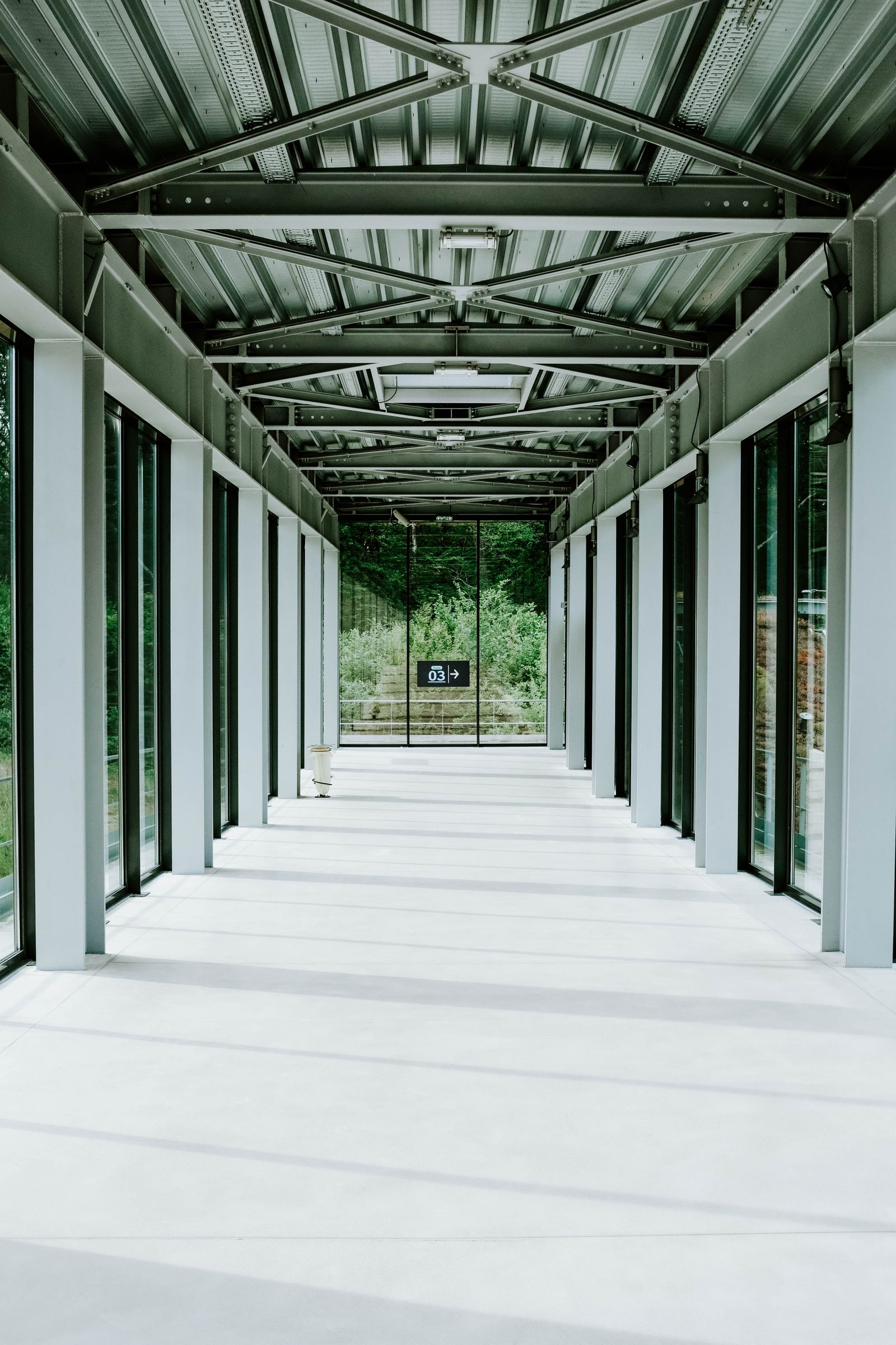 Long glazed corridor with repeated steel columns and roof trusses leading toward trees